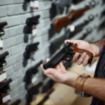 Person holding a handgun in a gun shop with various firearms displayed on the wall