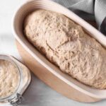 A loaf of bread dough resting in a wooden bowl next to a jar of sourdough starter