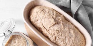 A loaf of bread dough resting in a wooden bowl next to a jar of sourdough starter