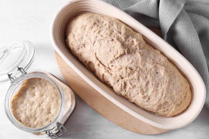 shutterstock_2456002407.jpg A loaf of bread dough resting in a wooden bowl next to a jar of sourdough starter
