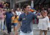 A man waving at a Pride parade with a rainbow flag pin on his shirt