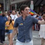 A man waving at a Pride parade with a rainbow flag pin on his shirt