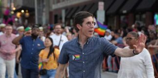 A man waving at a Pride parade with a rainbow flag pin on his shirt