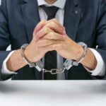 A man in a suit with hands handcuffed together on a table