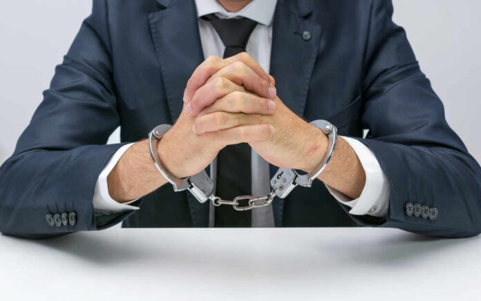 A man in a suit with hands handcuffed together on a table