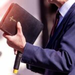 A person in formal attire holding a Bible while speaking