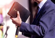 A person in formal attire holding a Bible while speaking