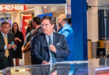 A news anchor speaking at a broadcast desk in a busy studio