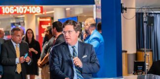 A news anchor speaking at a broadcast desk in a busy studio