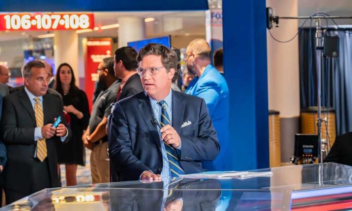 A news anchor speaking at a broadcast desk in a busy studio