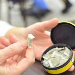 A hand holding a portion of snus with an open can in the background