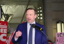 A man in a blue suit speaking at a protest with signs in the background