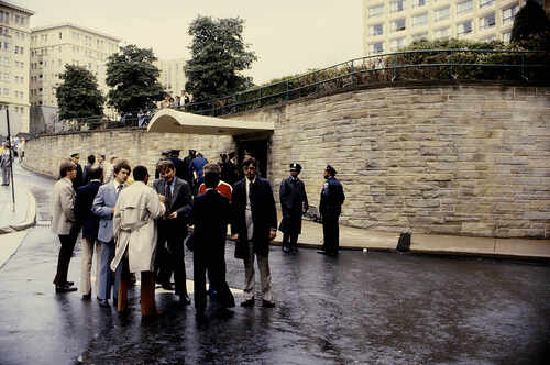 Group of people gathered outside a building with police officers nearby on a rainy day