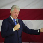 Man speaking with microphone in front of flag