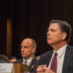 Government officials seated during a hearing with a nameplate in front of one