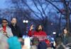 A woman in a red coat speaking at a podium during a rally, with audience members in the foreground