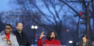 A woman in a red coat speaking at a podium during a rally, with audience members in the foreground