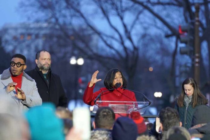 A woman in a red coat speaking at a podium during a rally, with audience members in the foreground