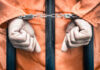 Hands of a prisoner in orange jumpsuit secured with handcuffs behind bars