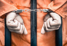 Hands of a prisoner in orange jumpsuit secured with handcuffs behind bars