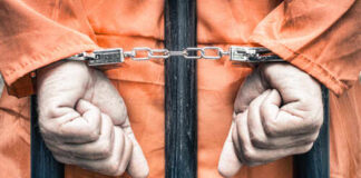 Hands of a prisoner in orange jumpsuit secured with handcuffs behind bars