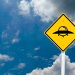 A yellow caution sign depicting a UFO against a blue sky with clouds