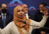 A woman in a hijab passionately speaking at a podium with a diverse group in the background