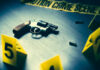 A handgun on a table with evidence markers and crime scene tape in the background