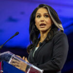 Cancer Battle Sparks Gabbard’s Shocking Departure Woman speaking at a podium with microphone
