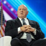 The Smear Failed—The Deportations Continued Man in suit sitting on stage with American flag.