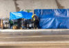 Two people at a sidewalk encampment with blue tarps and carts