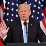 man speaking at podium with American flags behind
