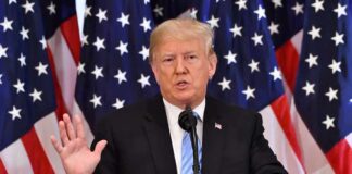 man speaking at podium with American flags behind
