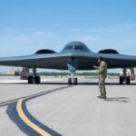 A military personnel stands in front of a B-2 Spirit stealth bomber on an airfield