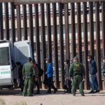 Trump’s ICE Crackdown Hits New Detention Record Border agents escort a group near a tall fence and a patrol van