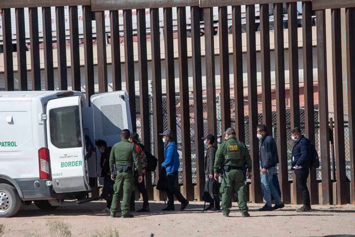 shutterstock_2079408475 Border agents escort a group near a tall fence and a patrol van