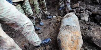Soldiers surround a large rusty unexploded bomb on rocky ground