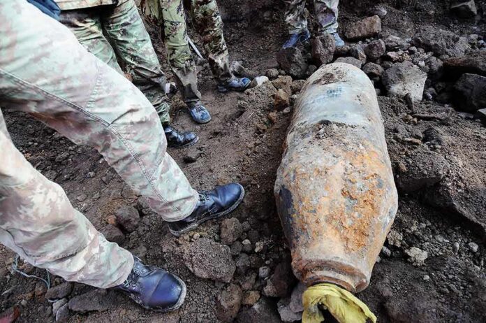shutterstock_1031166127 Soldiers surround a large rusty unexploded bomb on rocky ground