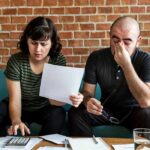 Middle Class Mirage: Mom And Dad Foot It Two adults on a sofa reviewing bills and using a calculator