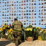 Double‑Tap Strike Slams Kyiv Rescuers Soldier kneels at memorial wall with photos and flowers