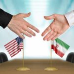 Two hands reaching to shake above US and Iran flags on a conference table