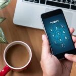 Hands holding smartphone with passcode screen near laptop and coffee
