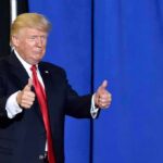 Donald Trump giving two thumbs up in front of a blue curtain