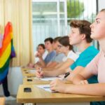 Students in classroom listening as teacher holds rainbow flag